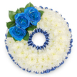 A bespoke memorial wreath shaped as the numeral '0', photographed from above against a clean white backdrop to emphasise its precise silhouette. The whole figure is meticulously covered in hundreds of tightly clustered white spray chrysanthemums, their tiny florets forming a plush, cloud-like surface that reads as softness and calm even at a glance. Around both the outer rim and the inner hole a pleated royal blue satin ribbon has been carefully laid - the ribbon's ruffled scallop echoes the circular form and frames the white chrysanthemums with a deep, refined contrast. At the upper left curve of the zero three royal blue roses are placed in a small, intimate cluster; their petals open from tight centers into satiny spirals, the deep sapphire tone striking against the white cushion beneath. Lush, dark green leaves nestle at the roses' bases, adding a quiet, natural depth and a hint of forest scent when the air is still. The arrangement is lit by soft, diffuse light that picks out the ribbon's sheen and casts gentle shadows between the chrysanthemum heads, giving the wreath a dignified, tactile presence. Crafted as a personalised tribute for a funeral or memorial, this number-shaped piece reads as both restrained and deeply considered - a way to mark an age or special date with colour and care. As a local florist serving Canonbury and nearby Islington, we often assemble such thoughtful numerical tributes for wakes at Canonbury Square churches or private homes along Essex Road, ensuring each bloom and fold of ribbon honours memory with quiet elegance.