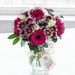 A vibrant mixed flower bouquet glows in soft daylight, set in a clear, textured glass vase and placed against a bright, gently blurred white background that feels like a sunlit Canonbury window. Deep fuchsia and magenta gerbera daisies with velvety dark centers form bold focal points, their wide petals curving outwards with cheerful confidence. Around them, rich purple-and-white chrysanthemums cluster together, each petal tipped in plum and fading toward creamy white near a golden-yellow eye, creating a tapestry of colour and texture. Creamy white roses, some fully open and others still in tight, elegant buds, nestle between the brighter blooms, lending a gentle, romantic softness. Fine sprays of airy white gypsophila thread through the arrangement, like tiny clouds catching the light, adding volume without heaviness. Lush, deep green foliage frames the flowers, its glossy leaves grounding the palette and hinting at the freshness of a garden walk through nearby Canonbury Square. The short, jar-style vase is cinched at the neck with a rustic twine bow, its fibres slightly frayed, suggesting a hand-finished touch. From this twine hangs a small, white rectangular gift tag, decorated with a simple pink floral emblem and neat lettering, ready for a personalised message for a birthday, anniversary, or just-because surprise. The clear water reveals neatly trimmed stems, carefully spiralled, reflecting the work of an experienced local florist used to preparing joyful deliveries for homes along Upper Street and the quiet terraces off St Paul's Road. The overall mood is one of bright, uplifting celebration, as if this bouquet has just arrived to bring colour, scent, and warmth into someone's living room.