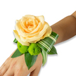 An intimate wrist corsage rests gently on the inner forearm, the skin a warm, light-to-medium tone that catches soft studio light, creating a calm, luminous backdrop for the flowers. At its heart sits a fully opened rose in a tender peach-apricot hue, its velvety petals layered in a slow spiral that invites a loving fingertip. Beneath the rose, two small lime-green button chrysanthemums-cheerful, almost spherical pom-poms-provide a lively, textural counterpoint; their tight florets and bright chlorophyll tone brighten the composition like a laugh beside a whisper. Deep, glossy leaves such as salal or ruscus form a dense, structured base, their rich green adding depth and a cool visual weight. A single variegated leaf, striped with soft cream and green and artfully folded into a sweeping arc, adds a modern, architectural flourish that guides the eye along the wrist and suggests motion when the wearer moves.
As a florist who often works in Canonbury and around Essex Road, I imagine this corsage worn at a local summer wedding at Canonbury Square or carried into a reception near Regent's Canal. The scent is a faint, elegant whisper of rose warmed by skin, underscored by a fresh grassy note from the chrysanthemums and foliage. The piece feels handcrafted, balanced between softness and structure-romantic yet contemporary-suitable for prom, an intimate ceremony, or a thoughtful honoured-guest gift. The fastening is discreet, likely an elastic or ribbon hidden beneath the leaves, leaving the floral artistry front and centre. Overall, this wristlet radiates warmth, celebration, and careful craftsmanship that complements both quiet mornings and evening festivities in Islington.
