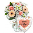 This image shows a joyful Mother's Day gift pairing arranged against a bright white background, where every element feels considered and calm. In the left half of the frame sits a clear glass vase, its surface smooth and transparent, revealing the stems within and finished at the neck with a casual tie of pale brown twine. From the vase rises a rounded bouquet in soft, uplifting tones. Fully opened peach-pink roses form the plush centre, their layered petals creating a sense of tenderness and care. Around them, several peach gerbera daisies with pronounced dark centres offer a playful, sunburst shape, echoing the kind of cheerful designs you might see crafted by Canonbury Flowers for local deliveries. Scattered throughout are fluffy clusters of white baby's breath, which lend a light, airy mist to the arrangement, and compact white chrysanthemums with subtle green hearts that add both texture and a fresh, garden-like feel. Deep and mid-green foliage is tucked between the blooms, providing a natural, leafy backdrop that helps each pastel tone stand out. Slightly behind the flowers, a small white card with a hint of colourful writing is nestled, its partial visibility suggesting a personal Mother's Day message. To the right, filling the remaining space, a large heart-shaped foil balloon leans towards the bouquet. The balloon is crisp white, sprinkled with reflective gold and silver confetti dots, and at its centre sits a peach heart inscribed with the words HAPPY Mother's DAY in bold black type and flowing script. Together, the flowers, card, and balloon create a scene that feels like a gentle celebration, ready to be delivered to a Canonbury home in honour of a much-loved mum.